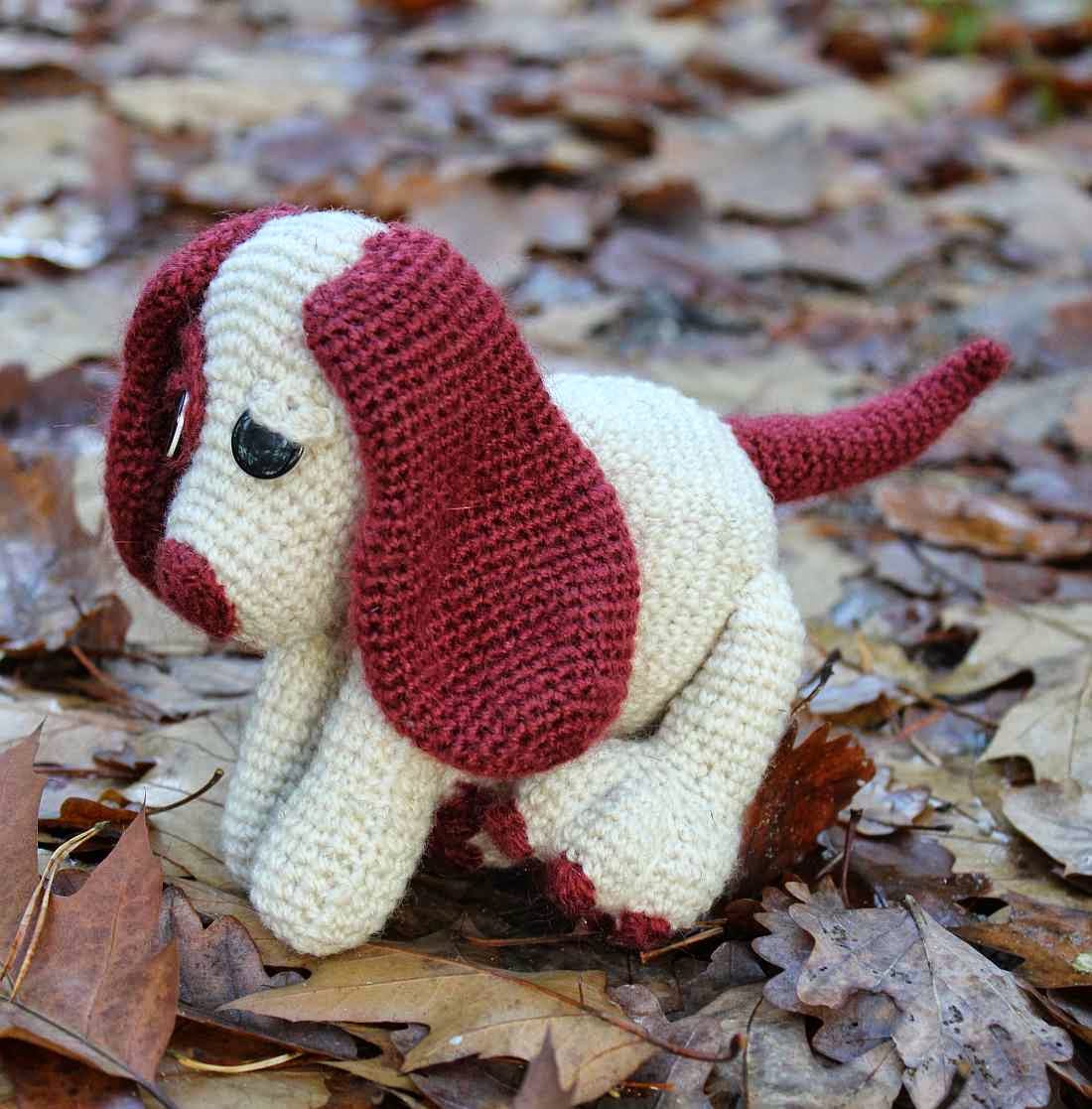 Crocheted cream and burgundy toy dog figure with floppy ears, sitting on a bed of dry autumn leaves, warm brown tones